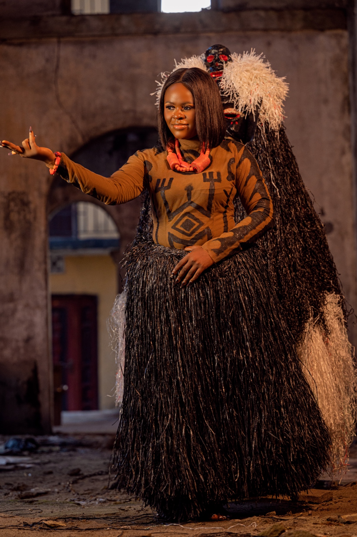 Ijaw woman in ochre body-paint tunic and coral itun beads standing beside the Blacky masquerade — black raffia column and carved wooden mask with pale tufted crown