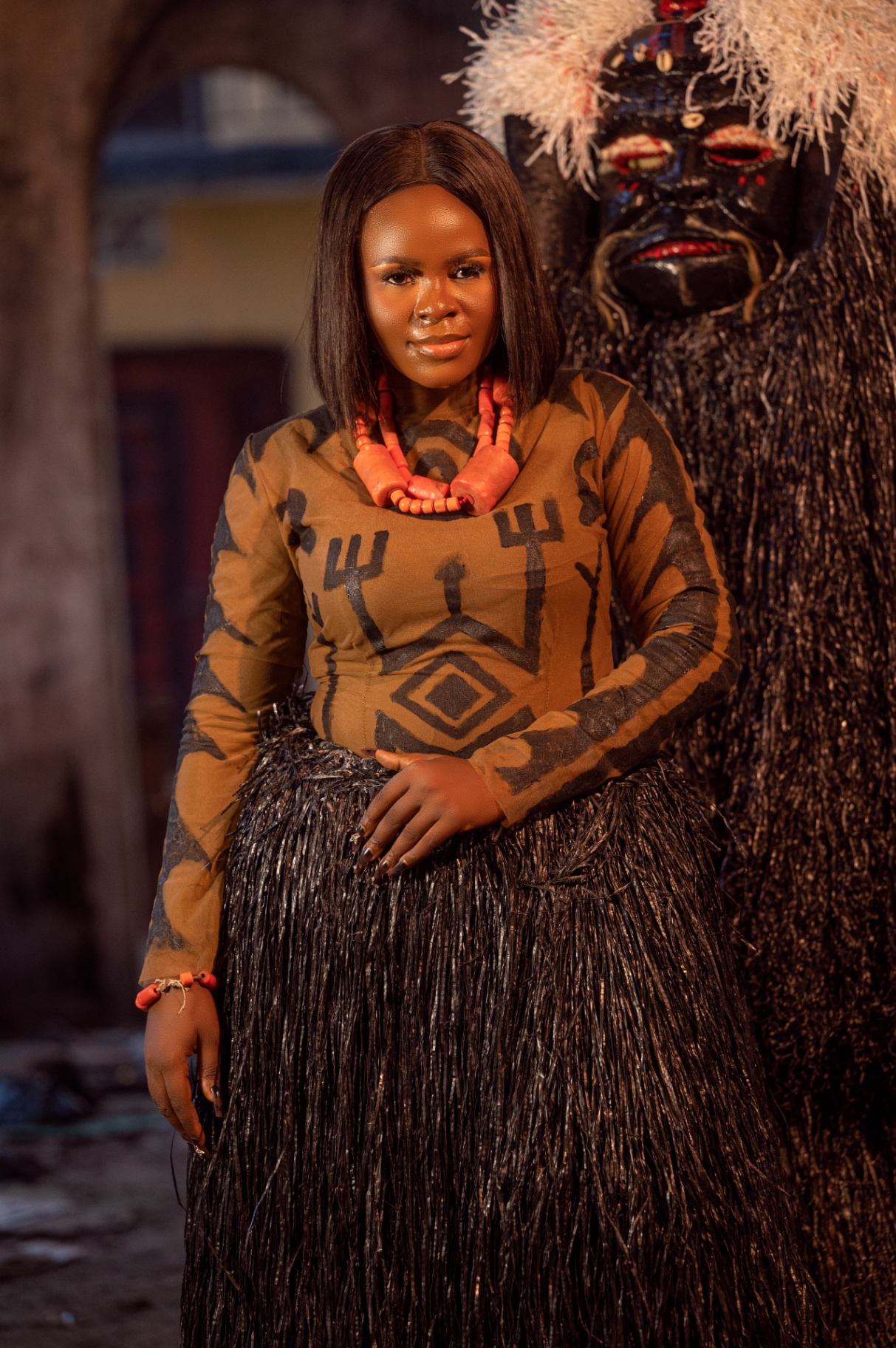 Portrait of the Blacky masquerade beside an Okrika woman in ochre body-paint tunic — carved black wooden mask with red lips, crimson orbital markings and pale tufted crown atop a dense black raffia column