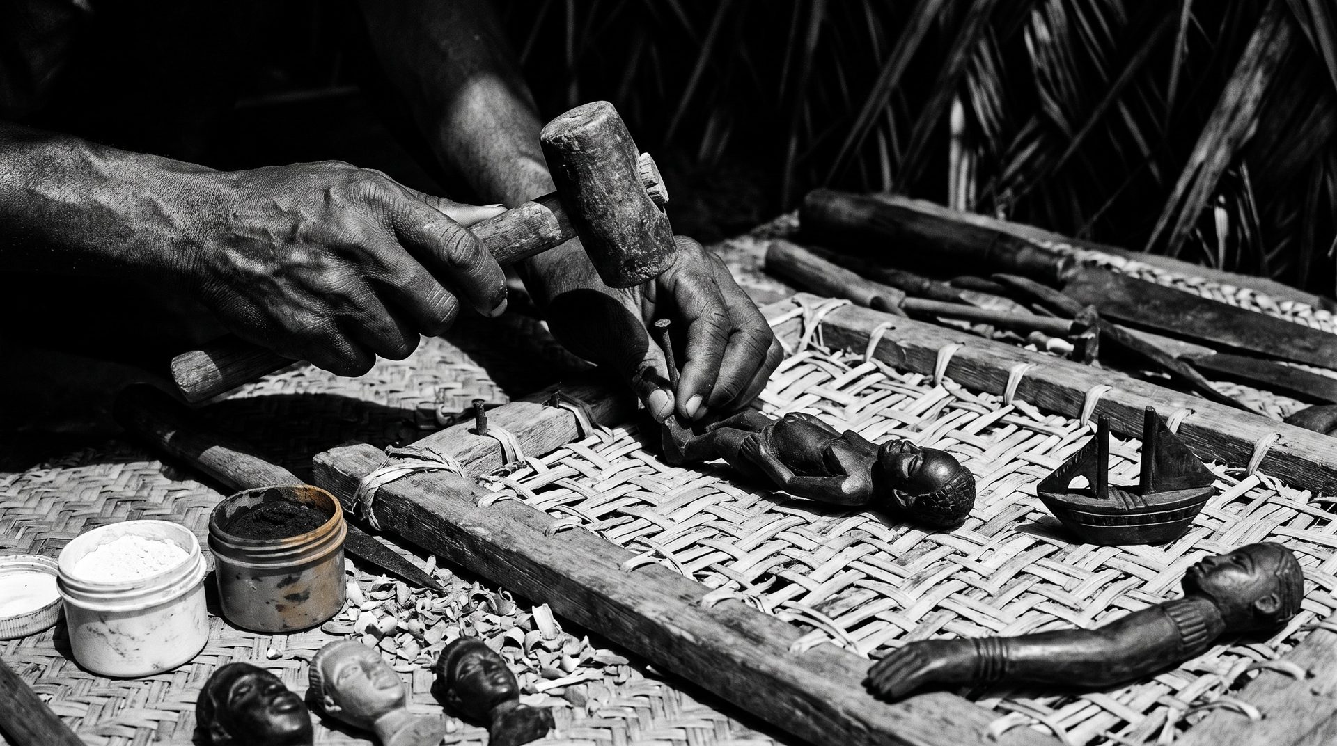 A woodcarver's hands fitting a separately carved wooden figure onto a cane backing inside a rectangular frame, with carved heads, a miniature ship headdress and pots of pigment laid out nearby