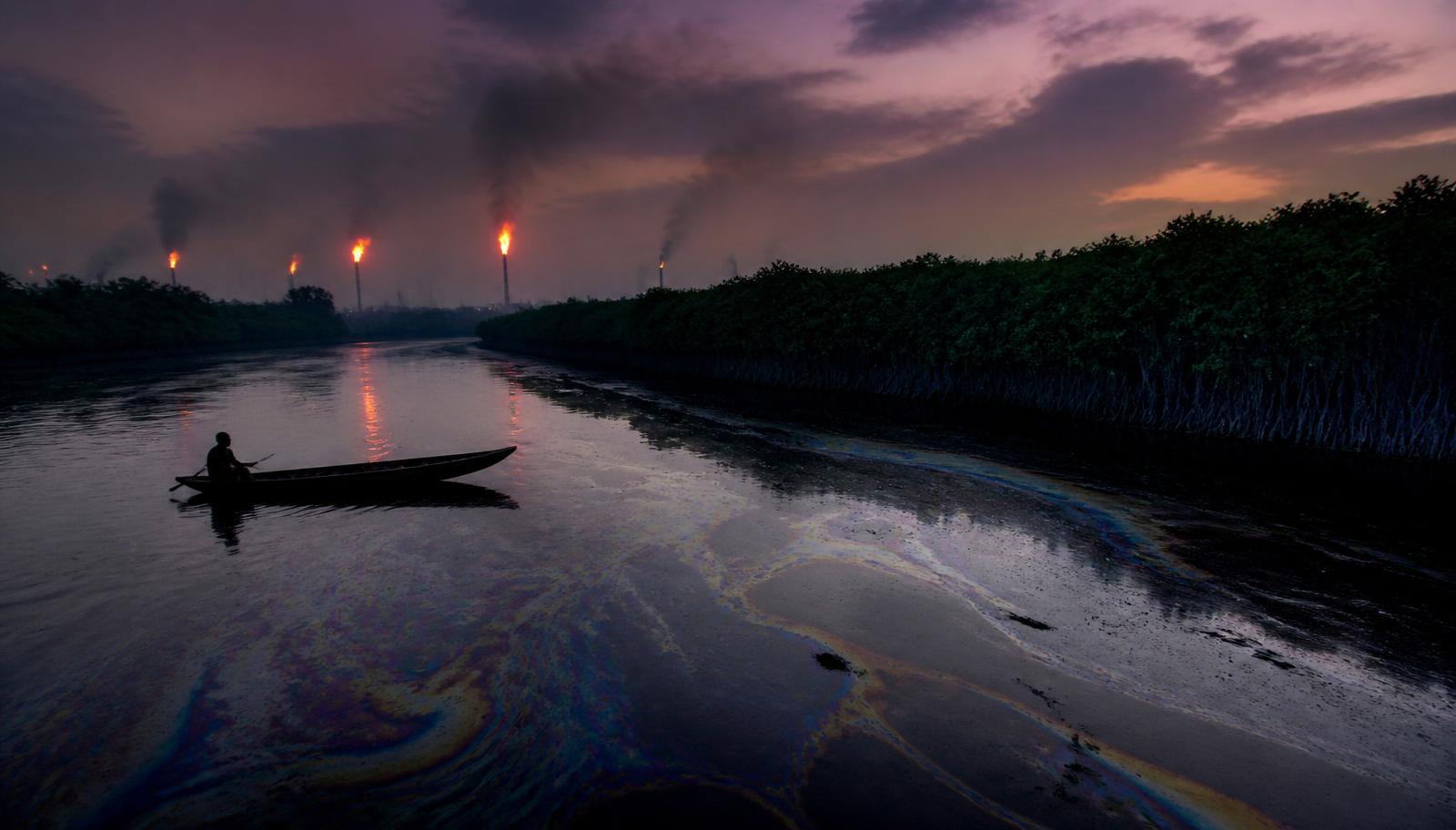 Niger Delta creek at dusk with oil sheen on the water and gas flares burning on the horizon