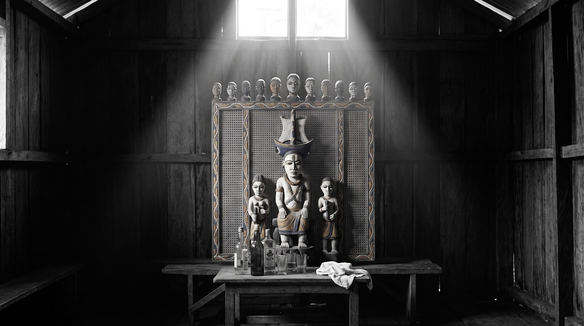 A carved Kalabari ancestral memorial screen in a dim meeting hall with an offering table before it