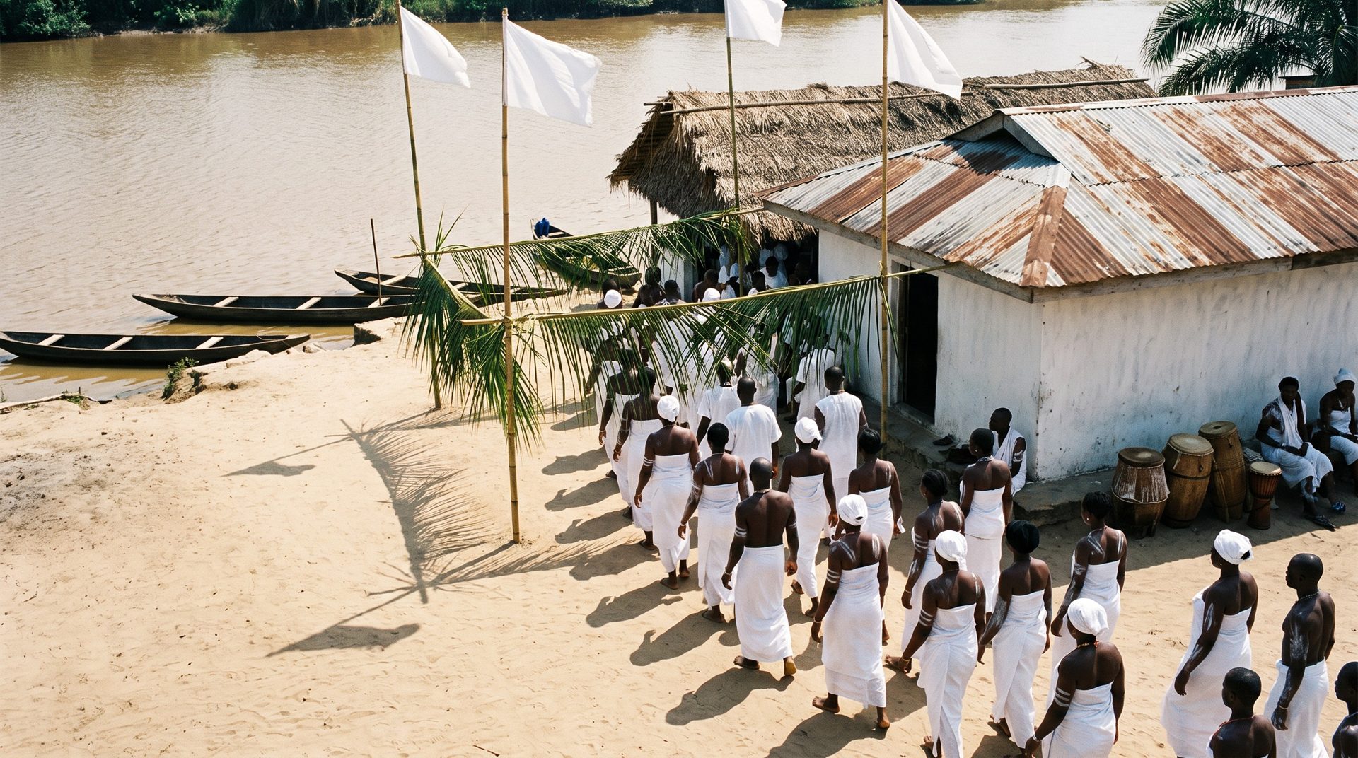 A whitewashed riverside shrine compound with tall bamboo poles flying plain white flags, devotees in white walking toward the temple, canoes moored on a brown creek