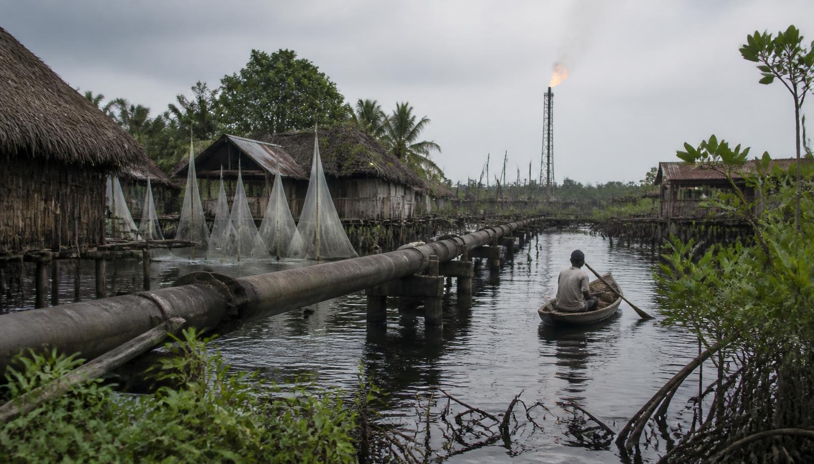 A trunkline running past stilt homes and fishing nets. The same water carries the canoe, the catch, the household supply and, when a line fails, the spill.